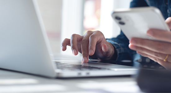 Close-up of a person using a laptop while holding a smartphone