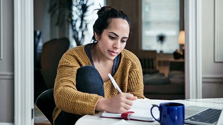 Person writing in a notebook at a table beside a laptop and coffee mug