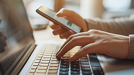Close-up of a person typing on a laptop while holding a smartphone