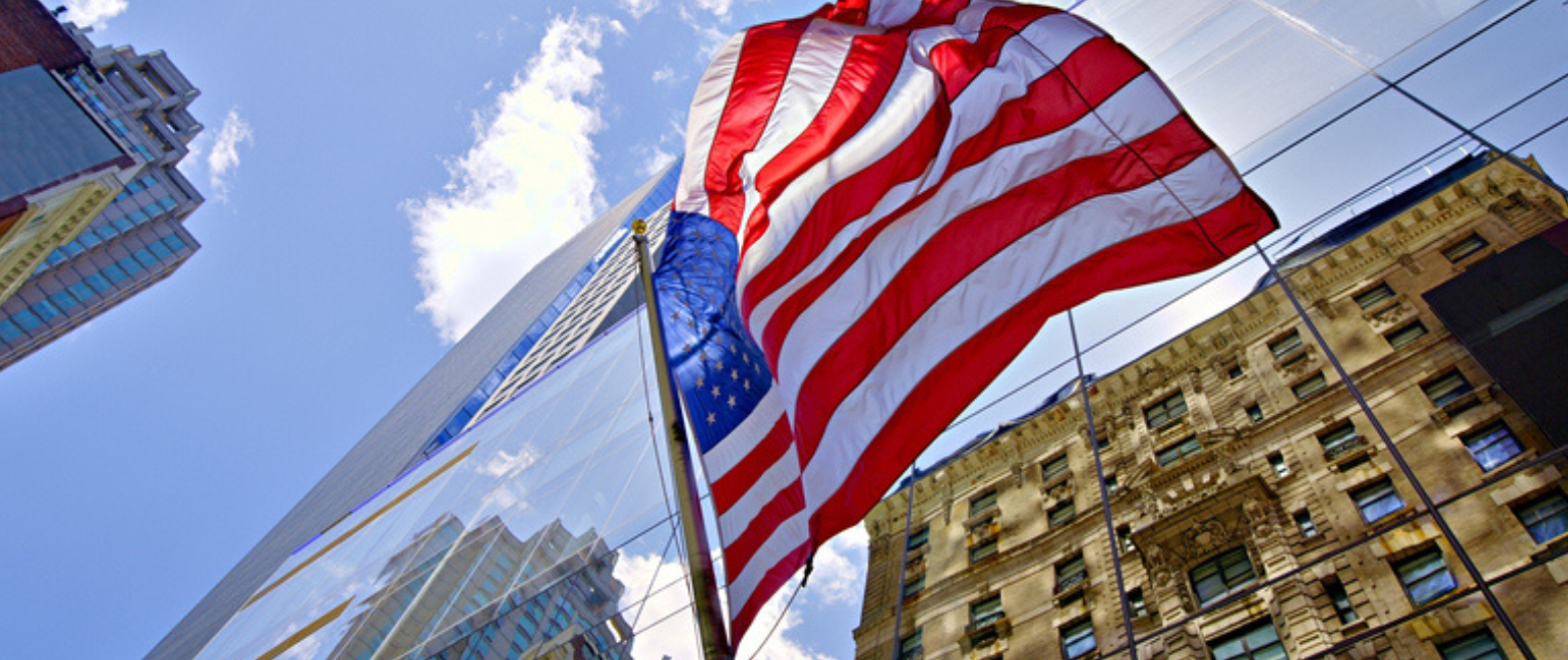American flag waving against city skyscrapers and a blue sky.